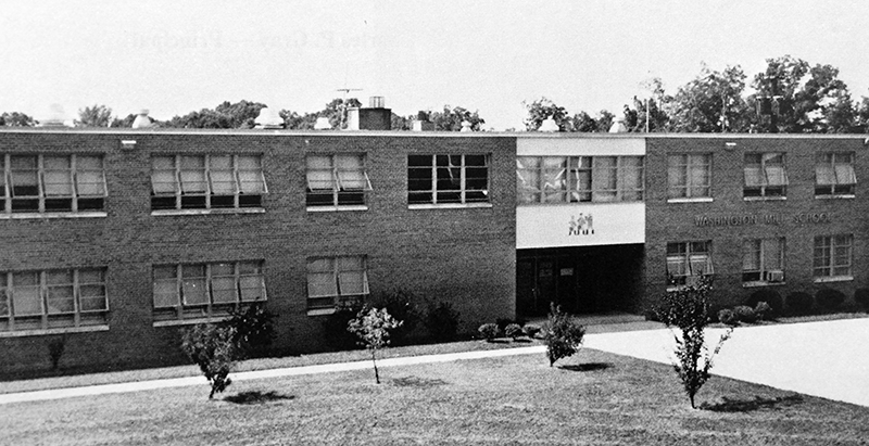 Black and white yearbook photograph of Washington Mill Elementary School taken in 1972. The trees and shrubs on the school grounds are still quite small. The classroom windows on both floors of the building are propped open. 