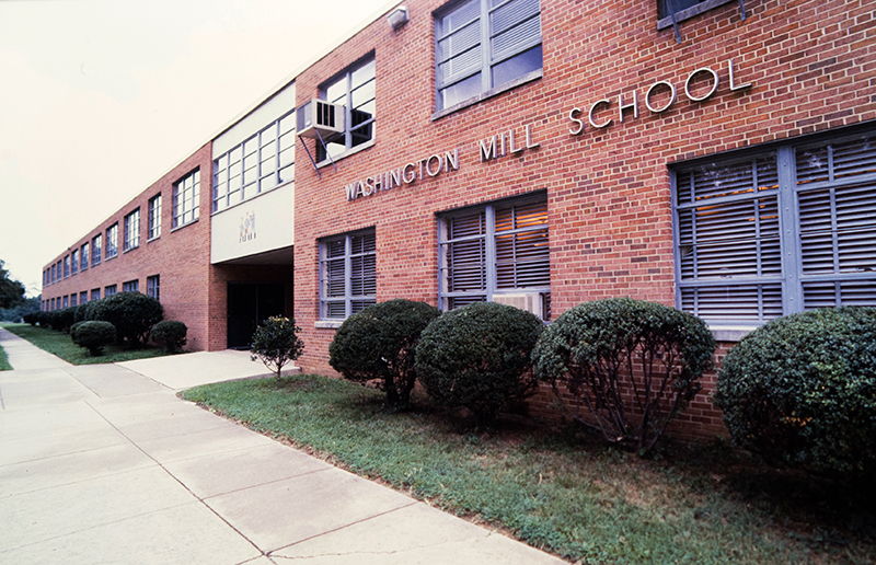 Color photograph of Washington Mill Elementary School's main entrance taken between 1983 and 1988. The windows are closed and two rooms have window-mounted air conditioning units installed. 