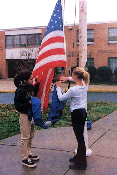 Color photograph from our 1997 to 1998 yearbook showing two students raising the American and Virginia flags on the pole in front of our school. 