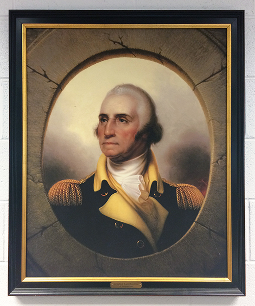 Photograph of the Washington portrait on the wall in our library. It is a portrait of Washington in later life when he was President of the United States. He is wearing a gold and black or dark blue uniform with gold epaulettes. There is a gray oval matte around Washington and the picture is hung in a black and gold bordered frame. 